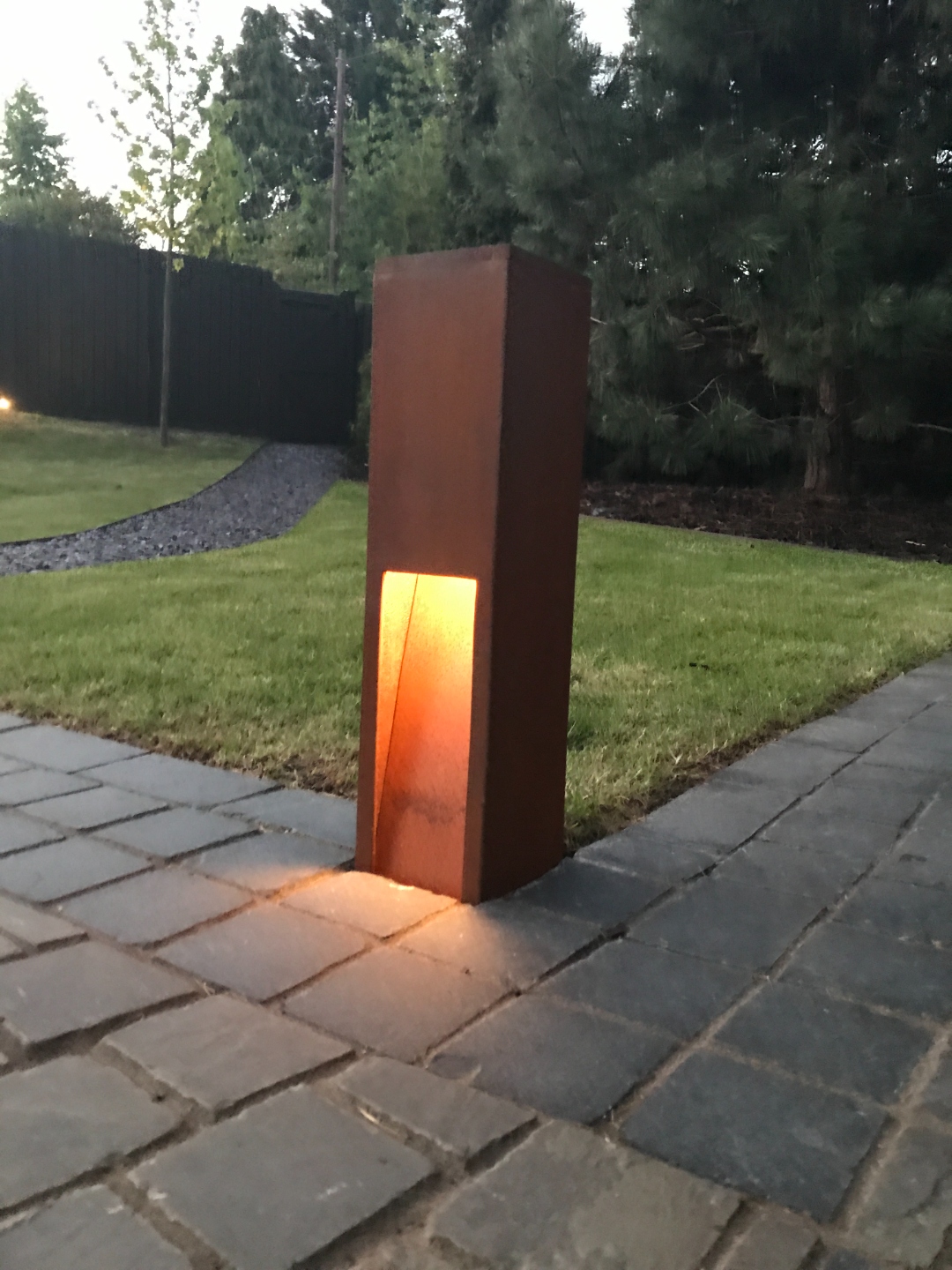 Corten steel garden light bollard on block-paved path at dusk